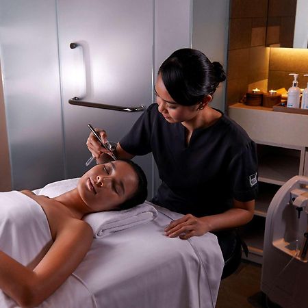 Paradox Singapore Merchant Court At Clarke Quay Εξωτερικό φωτογραφία The photo shows a spa or beauty therapy setting where a woman is lying on a treatment table with a towel draped over her. A professional, dressed in a dark uniform, is performing a skincare treatment on her face. Various beauty products are visible o