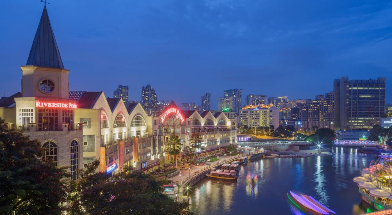 Paradox Singapore Merchant Court At Clarke Quay Εξωτερικό φωτογραφία The photo shows a riverside scene at dusk, featuring a waterfront area with modern buildings that have a distinctive architectural style. The structures have pointed roofs and are well-lit, creating a vibrant atmosphere. Along the river, there are bo