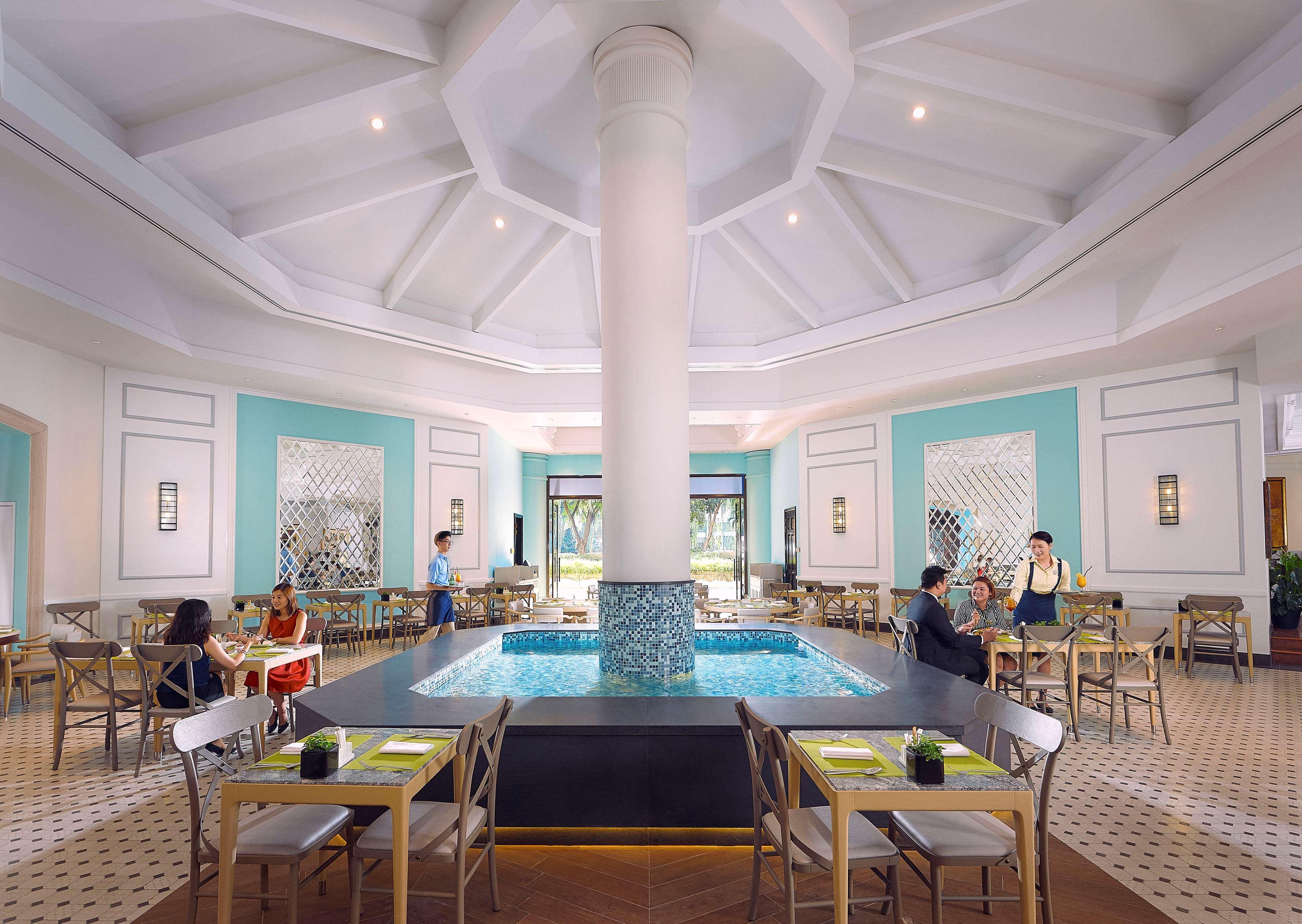 Paradox Singapore Merchant Court At Clarke Quay Εξωτερικό φωτογραφία The photo depicts an elegant restaurant interior characterized by a spacious, well-lit design. At the center, there is a large, round column that supports a high, intricately designed ceiling. Surrounding the column is a small pool of water, adding a