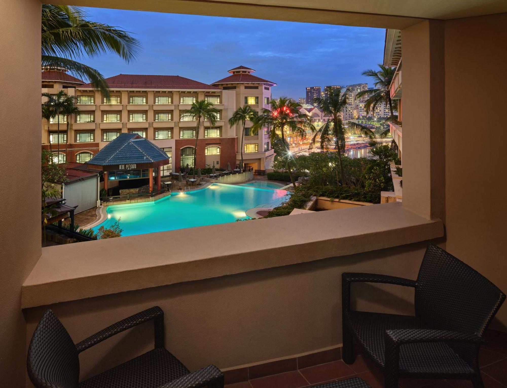Paradox Singapore Merchant Court At Clarke Quay Εξωτερικό φωτογραφία The image depicts a view from a balcony overlooking a pool area of a hotel or resort. The scene features palm trees surrounding the pool, which has a blue hue, possibly illuminated by lights. In the background, there are several buildings, likely par